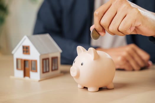 A businessman hand saving money by placing a coin into a piggy bank next to a house model, representing investment, financial stability, property goals, and a savings-focused approach to home buying.