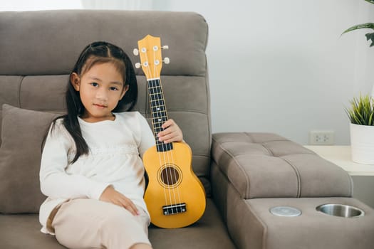 A young girl in a white shirt sits on a grey couch, holding a small wooden ukulele. Her bright smile and direct gaze at the camera radiate warmth, cheerful and creative moment in a cozy living room.