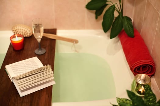 Time for Yourself. Relax at home. Bath tub with flower petals. Book, candles and glass of wine on a wood tray. Organic Spa Relaxation in comfort cozy bathroom