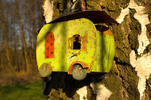 A yellow birdhouse in the shape of a caravan, attached to a tree trunk with white bark, in the sunlight. Ideal for nature and garden lovers.