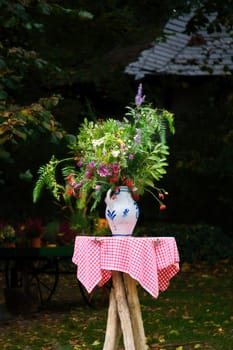 A bouquet of flowers in a vase on a table with a red and white checkered cloth, in the background a garden with trees and plants. Ideal for nature and garden lovers.