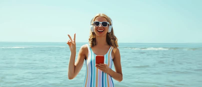 Summer vacation of happy young woman smiles enjoying listening to music with headphones, holding phone, relaxed cheerful girl wear a sunglasses on beach at sea coast with blue sky on a sunny day