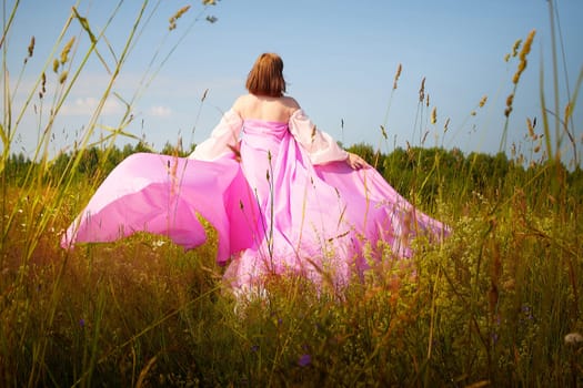 A woman wearing a flowing pink dress stands amidst tall grasses and colorful wildflowers on a sunny day, embracing nature's beauty.