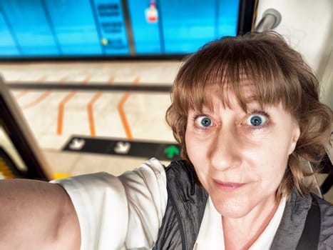 A middle-aged woman with blonde hair captures a joyful selfie at a bustling train station, excited for her upcoming journey.