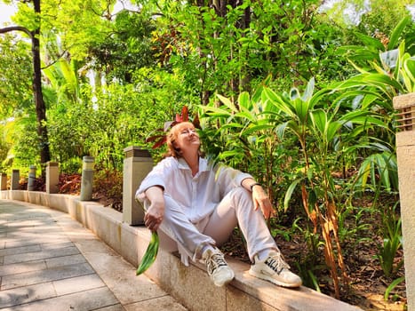 Blonde woman sits peacefully on a stone ledge surrounded by vibrant greenery, enjoying her travel adventure and the beauty of nature.