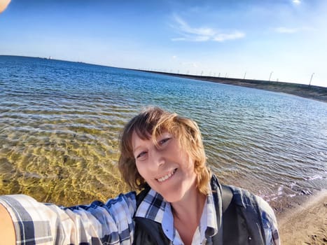 Smiling blonde woman takes a selfie by the beach, enjoying a warm day by the water and capturing her travel adventure.