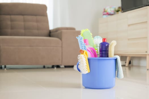 Cleaning tools and supplies neatly stored in blue bucket. Detergent bottles, sponges, and brushes sit on floor in living room, making housework and daily cleaning tasks more convenient and organized.