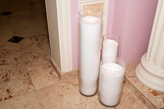 Three large candles stand on the marble floor. Candles in glass vases against a pink wall.