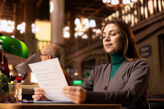 Focused senior woman studying at a wooden desk in a public library, using textbooks and academic archived files to symbolize the lifestyle of adult education and intellectual pursuit.