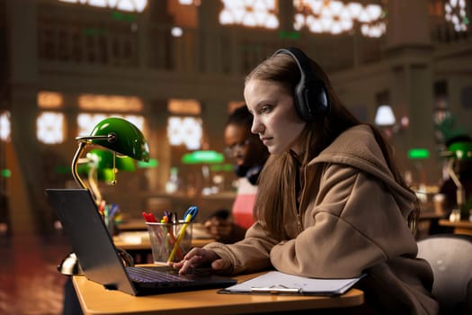 University scholar listening to therapy music on headset to create the perfect ambient for research, working on her diploma paper. Young girl uses focus music to help with collecting information.