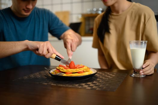 Happy young couple enjoying pancakes with strawberries and syrup in cozy home kitchen.