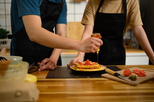 Close-up of man pouring honey over stacked pancakes with strawberries in a cozy kitchen..