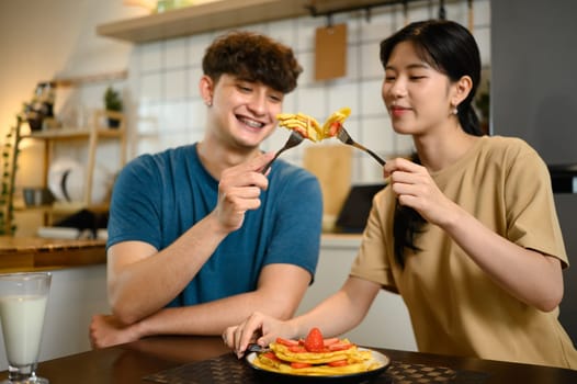 Happy young couple enjoying pancakes with strawberries and syrup in cozy home kitchen.