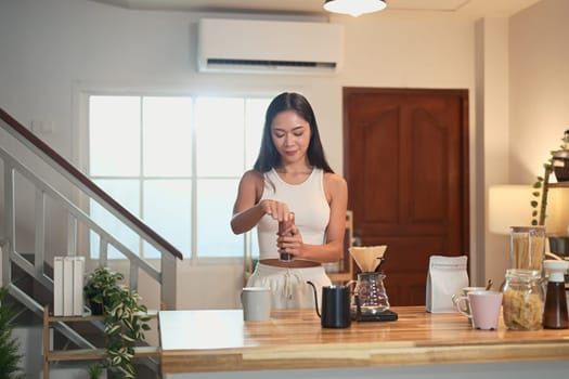 Young woman grinding coffee beans and preparing pour over coffee in a warm kitchen.