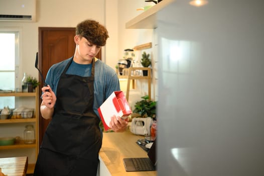 Young man in apron reading pancake mix instructions in a cozy kitchen.