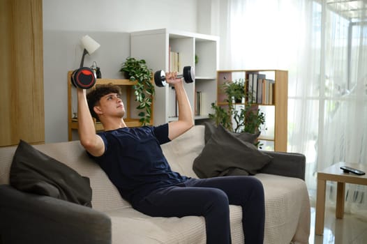 Young man lifting dumbbells on a sofa. Fitness at home concept.
