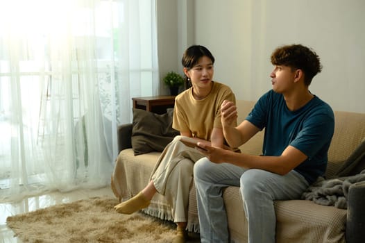 Young couple sitting on sofa, having a conversation and using smartphone in living room.