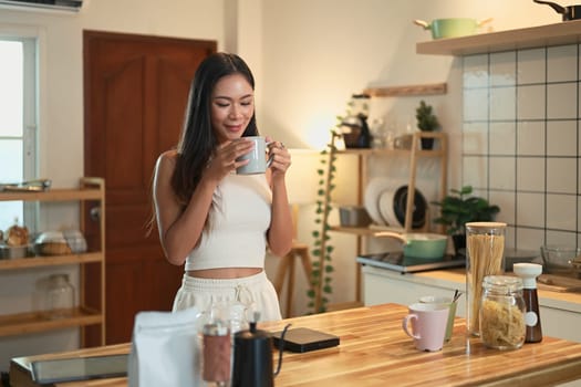 Smiling woman savoring a warm cup of coffee while standing in a cozy, modern kitchen.