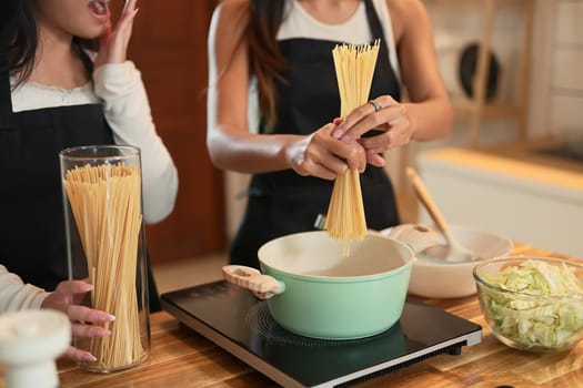 Mother breaking dry spaghetti over a pot while her teenage daughter reacts with surprise.