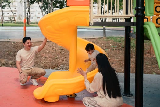 Happy parents playing with their young child at the playground slide on a sunny day..