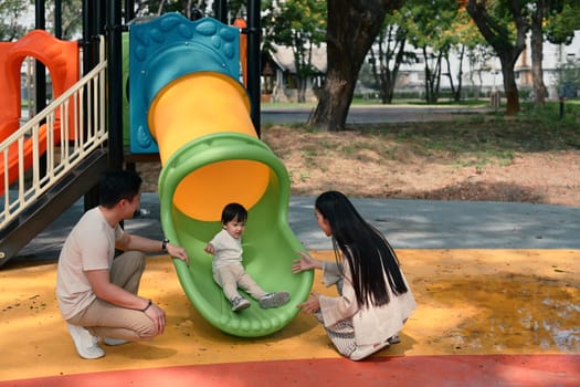 Happy parents playing with their young child at the playground slide on a sunny day..