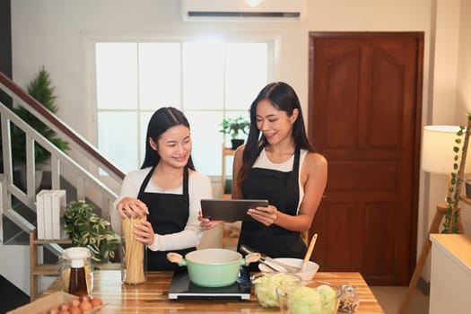 Asian mom and teenage daughter reading digital recipe together while preparing a home cooked meal.