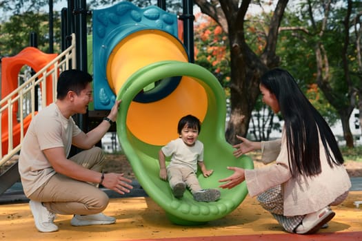 Happy parents playing with their toddler on a colorful playground slide at the park.