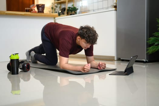 Young man doing plank exercise at home while following a workout video on a tablet.