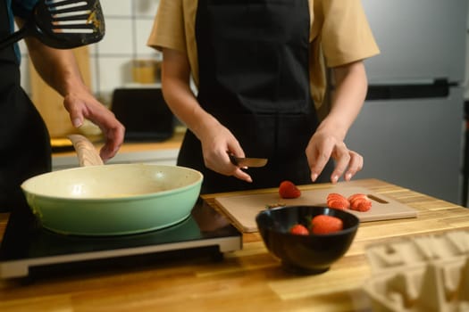 Close up of couple preparing pancakes, slicing strawberries and cooking on stovetop.