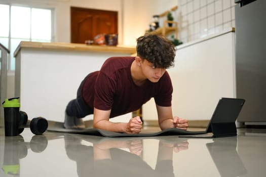 Young man doing plank exercise at home while following a workout video on a tablet.