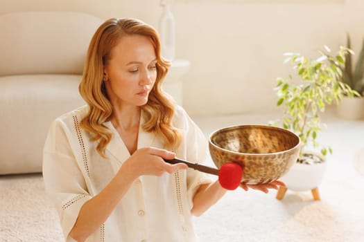Calm woman using Tibetan singing bowl for sound therapy and meditation in a peaceful home interior with soft light.