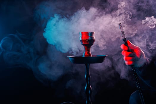 Hookah with dense smoke and vibrant red-blue lighting, hand holding pipe hose in dark moody background.