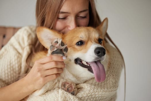 Smiling woman cuddling happy Corgi dog, holding paw up, close bonding moment showing love and affection indoors.