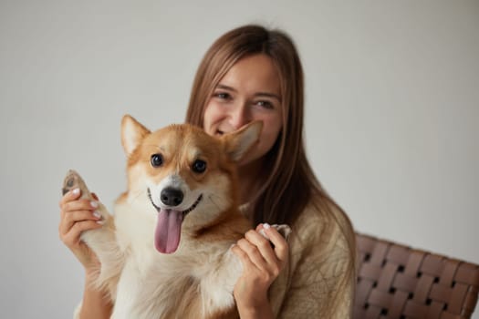 Cheerful woman holding a happy Corgi dog raising its paws, joyful friendship moment, cozy home vibe, neutral background.
