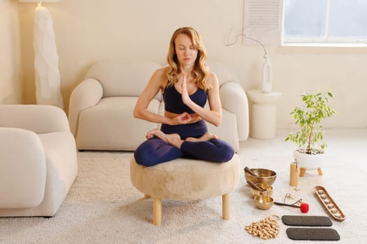 Woman in yoga outfit meditating on soft stool in zen space with healing instruments, candles, and nail boards.