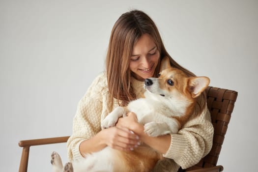 Smiling woman cuddling a cute Corgi dog on a chair, cozy winter moment, friendship and love, on neutral background.