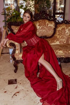 young and luxurious girl sits on a golden empire style sofa. girl in a red long evening dress. silk dress has long sleeves. girl has a high hairstyle and evening make-up. she has long legs and sandals