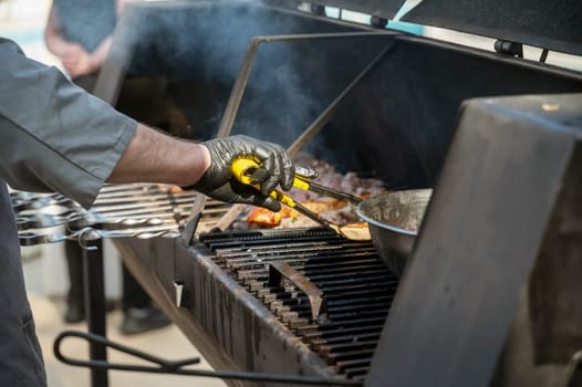 Male chef preparing meat on grill at barbecue party in summer garden, chef's hands in gloves. Concept of food, people and family time. Pork on skewers is roasted on coals in large metal grill device