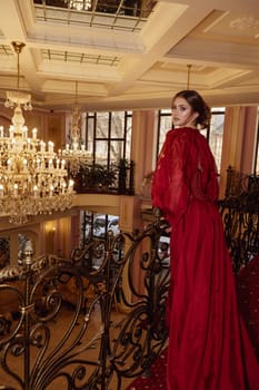 model in a red long dress against the background of a luxurious hotel interior. interior with crystal chandeliers, mirrors, forged elements in the interior. elegant young woman in the hotel lobby.