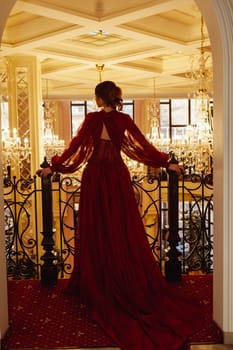 Silhouette of a model in a red dress against the backdrop of a luxurious hotel interior with crystal chandeliers. View from the back of an elegant young woman in a long evening dress