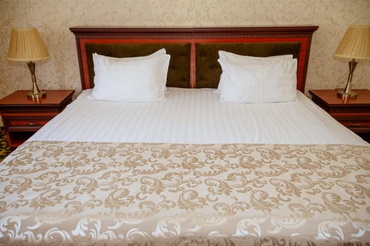 Double bed with wooden, dark red headboard, clean white bed linen and pillows. bedspread and wallpaper of the same golden beige color. On the bedside tables are table lamps with golden lampshades