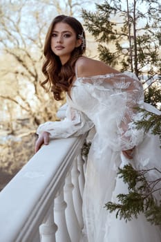 Young brunette bride in traditional long dress posing on white hotel balcony in spring. white dress with long sleeves, corset and open shoulders. Bride portrait, wedding makeup, fashionable bride.