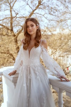 Young brunette bride in traditional long white dress posing on hotel balcony in spring. Long white dress with long sleeves, corset and open shoulders. Bride portrait, wedding makeup