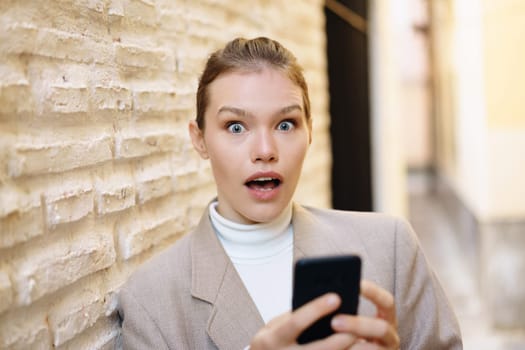 An astonished woman, displaying a shocked expression, is intently holding her smartphone, with a textured wall behind her, situated in a lively urban setting that captures the essence of modern life
