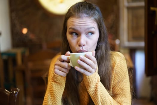 A young woman in a cozy caf sips coffee, lost in thought. She wears a warm sweater, embodying a moment of relaxation and introspection
