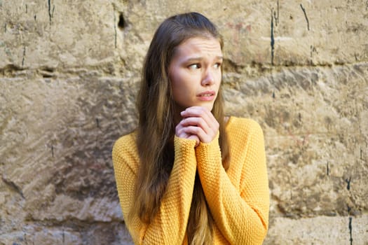 A young woman clad in a cozy yellow sweater stands against a beautifully textured stone wall. Her expression reflects deep worries, evoking strong emotions of concern, unease, and anxiety in viewers