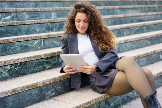 A stylish young woman with beautiful curly hair sits comfortably on elegant marble steps, fully engaged with her tablet device, effortlessly embodying the modern worklife balance concept
