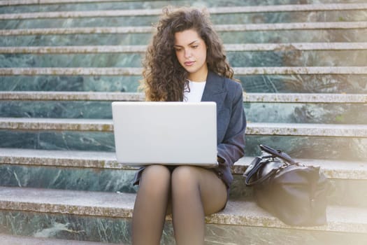 A young professional with curly hair is sitting on outdoor steps, focused on her laptop. She is dressed smartly, with a stylish bag beside her, embodying productivity and modern work culture.