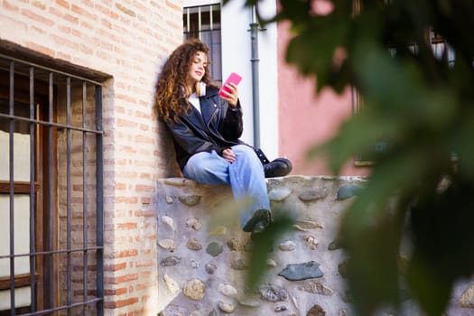 In a busy urban setting, a young woman with curly hair sits on a stone wall, focused on her smartphone, surrounded by vibrant green foliage, blending modern life with natures tranquility