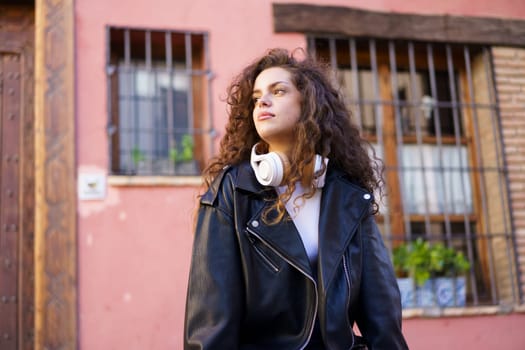 A fashionable young woman with beautiful, curly hair confidently poses against a vibrant and colorful wall, showcasing her trendy leather jacket and stylish headphones in a lively urban environment